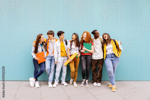 Happy multiracial students standing over isolated blue background. Diverse teenage friends having fun talking leaning on campus building college wall