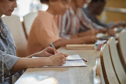 Close-up of schoolgirl sitting at desk and making notes during lecture with her classmates sitting in a row at desk