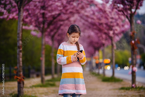 Little preschooler girl standing in striped dress before of blooming pink sakura alley