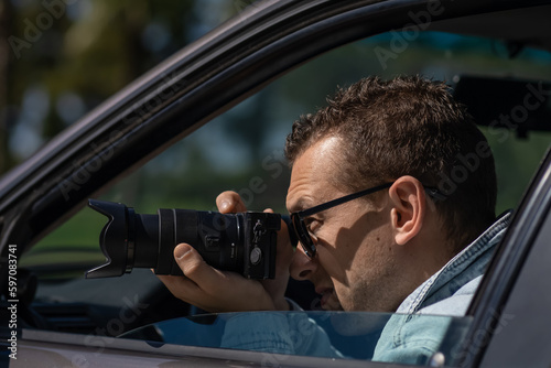 Man in sunglasses with camera sits inside car and takes pictures with professional camera, private detective or paparazzi spy. Journalist seeks sensation and follows celebrities.