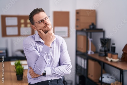 Young hispanic man at the office with hand on chin thinking about question, pensive expression. smiling with thoughtful face. doubt concept.
