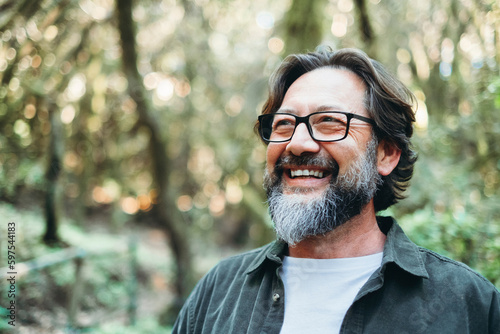 Portrait of young mature man alone smiling outside with nature trees in background. Environment. Happy people enjoying outdoor leisure activity. Nature feeling. Bearded adult male wearing eyeglasses