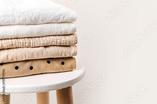 Stack of baby clothes. Cotton clothes and muslin swaddle blanket in white and beige colors. Clean freshly laundered, neatly folded kids clothes on table.