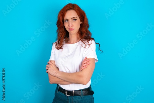 Displeased young redhead woman wearing white T-shirt over blue background with bad attitude, arms crossed looking sideways. Negative human emotion facial expression feelings.