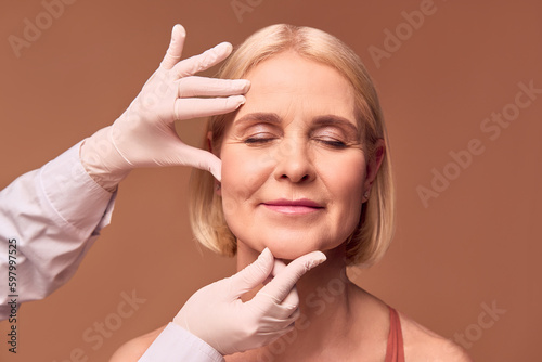 Portrait of an older adult woman with closed eyes on a beige background. Hands in white gloves and a medical gown show a face that has wrinkles.Face lift.
