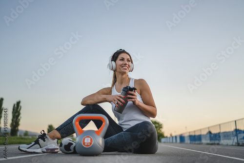 one caucasian woman female athlete rest hold supplement shaker kettlebell girya weight while sitting in stadium on track during training in summer evening copy space health and fitness concept