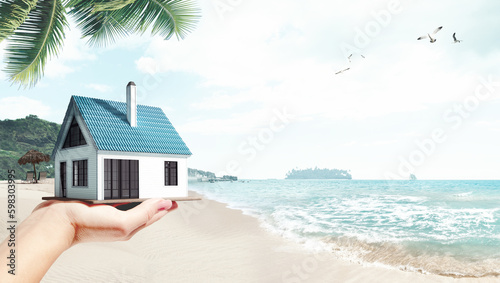 Moving to island. Female hand holding 3D model of small comfortable house against beach, palms and ocean background. Concept of real estate, buying house, mortgage, ownership, business