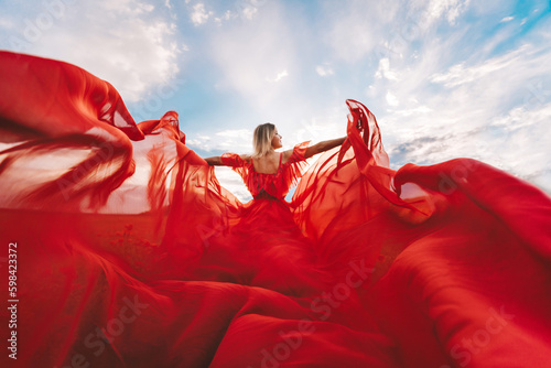 Woman Red Flying Dress. A blonde in a red dress against the sky. Rear view of a beautiful blonde woman in a red dress fluttering in the wind against a blue sky and clouds.