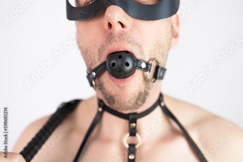 Muscular man in mask wearing leather harness and ball gag on white background.