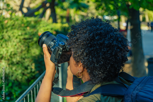 Young black African American woman with dark curly hair photojournalist taking a picture in a green park adjusting the focus ring and parameters of the photo camera