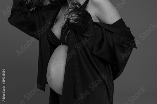 A pregnant woman in a black suit on a gray background. A pregnant woman. Studio pregnancy photo shoot. Stylish pregnancy. Black and white photo. Pregnant model posing in black dress