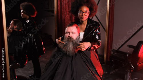 an African-American hairdresser girl straightens her beard with her hands to a male client sitting in an armchair.