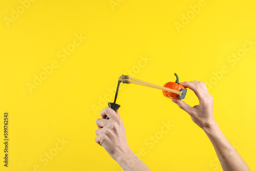 Hands holding slingshot with toy pumpkin on a yellow background. Halloween theme, trick or treat