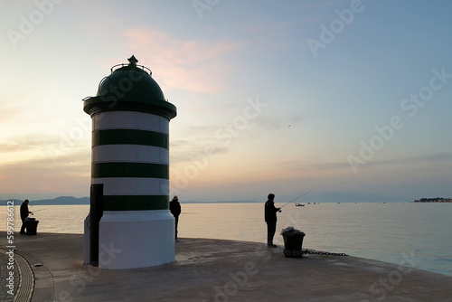 silhouette of people standing on pier fishing next to lighthouse against sunset sky in Zadar, Croatia