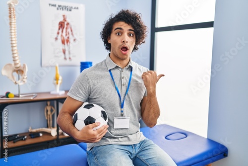 Hispanic man with curly hair working as football physiotherapist surprised pointing with hand finger to the side, open mouth amazed expression.