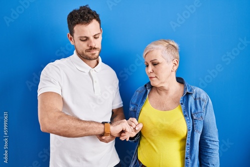Young brazilian mother and son standing over blue background checking the time on wrist watch, relaxed and confident