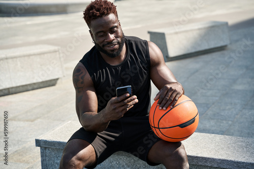 Fit sporty young African black ethnic man sitting outdoors holding basketball ball and mobile phone using apps, looking at smartphone, resting after street sport game with cellphone.
