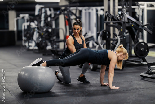 A female coach is training a fit sportswoman who is doing exercises with fitness ball.