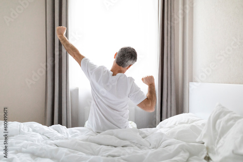 Back view of grey-haired man in pajamas stretching in bed
