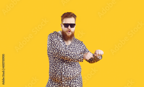 Plus size male model in funny pajamas having fun in studio. Bearded fat man wearing cool sunglasses and comfortable leopard PJs dancing isolated on yellow background. Fashion and crazy party concept