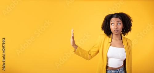 Serious african american lady doing stop gesture with hand and looking aside at free space, yellow background, panorama