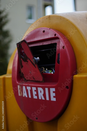 Zbliżenie na uchylone drzwi zbiornika na zużyte baterie wbudowanego w większy, żółty śmietnik