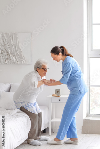 Young caregiver helping senior woman with stick to stand up in bedroom