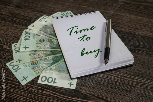 notatnik z wiadomością: "time to buy". Obok notesu na blacie stołu rozłożone banknoty.
