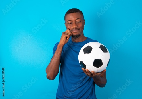 Pleased Young man wearing blue T-shirt holding a ball over blue background with closed eyes keeps hands near cheeks and smiles tenderly imagines something very pleasant