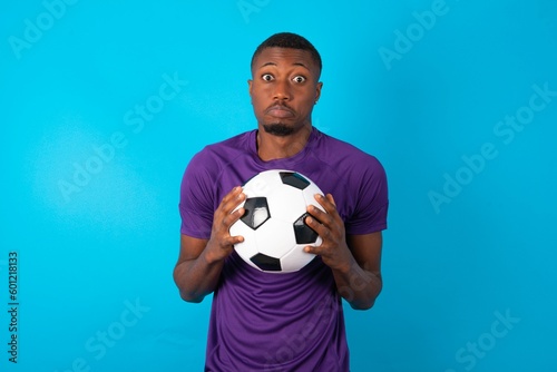 Happy Man wearing purple T-shirt holding a ball over blue background stands against orange studio wall keeps hands on heart, swears be loyal, expresses gratitude. Honesty concept.