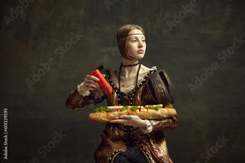 Portrait of young girl, queen, princess in vintage costume putting ketchup on giant sandwich baguette on dark green background. Concept of history, renaissance art, comparison of eras, health and food