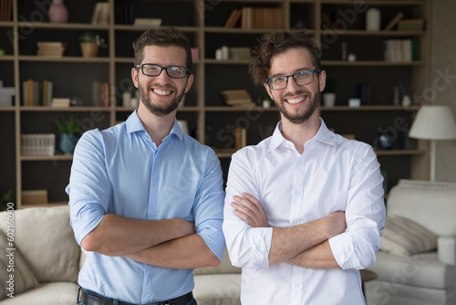 Positive confident family business partner men portrait. Happy handsome young adult twin brothers in glasses and office shirts posing together at home with arms crossed, looking at camera, smiling