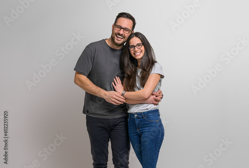 Portrait of loving young couple embracing and holding hands while posing ecstatically against background. Charming girlfriend and boyfriend wearing eyeglasses standing together and looking at camera