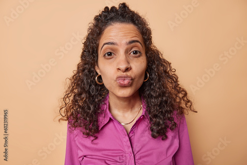 Attractive flirty young woman folds lips in mwah gives kiss at camera wears stylish purple shirt has romantic expression poses against brown background. Tender attitude and love confessing concept.