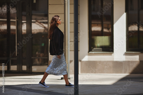 Beautiful brunette woman walks street city near buildings, wears luxurious attire clothes shiny maxi skirt with sequins, black sneakers and Tshirt. Light and shadow