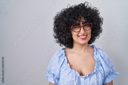 Young brunette woman with curly hair wearing glasses over isolated background winking looking at the camera with sexy expression, cheerful and happy face.