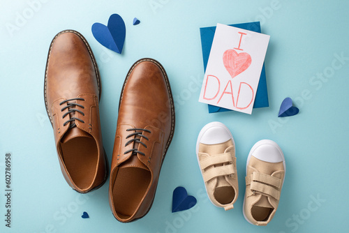 Baby boy surprises dad with heartfelt Father's Day gifts. Top-down view shot of dad's leather shoes, son's sneakers, hearts, handmade postcard, and envelope on pastel blue background
