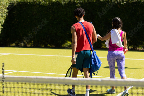 Rear view of diverse couple with bags and rackets walking on sunny outdoor tennis court, copy space