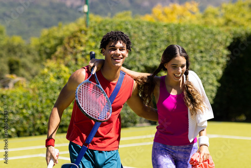 Happy diverse couple with bags and rackets embracing on sunny outdoor tennis court