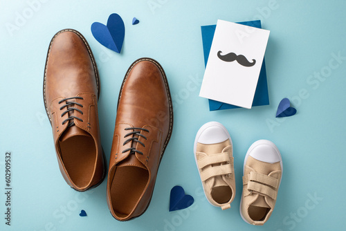 Little son shows love for dad on Father's Day. View from above of dad's leather shoes, baby boy's sneakers, heart embellishments, and postcard with mustaches on pastel blue background