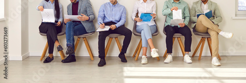 Legs of unrecognizable business people sitting on the chairs in a row with resumes or documents in their hands. Group of a staff. Job candidates seekers waiting for interview invitation turn. Banner.