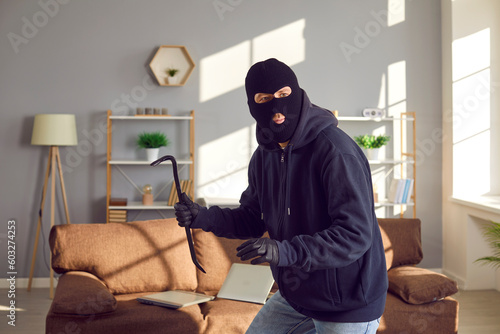 Burglar, robber, thief breaks into somebody's home. Man wearing black balaclava and gloves holding crowbar, walking on tiptoes and looking around for things he can take from this house or apartment
