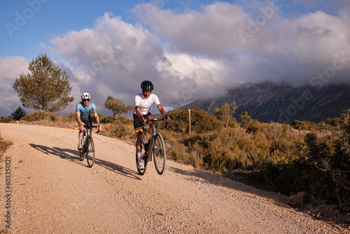 Man cyclist on gravel bicycle riding wheelie on a rear wheel.Two cyclists riding together on gravel path in spanish mountain at sunset