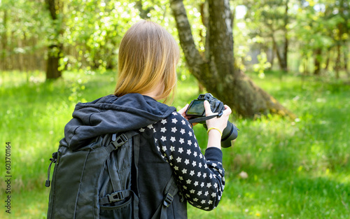 Kobieta z aparatem fotograficznym na szyi ogląda zrobione zdjęcia na ekranie lustrzanki stojac w lesie 