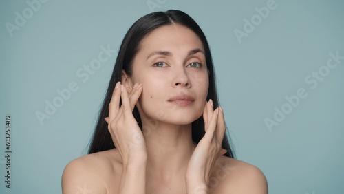 A seminude woman touches her clean and smooth face with wellgroomed fingers. Close up portrait of young woman isolated on blue background. Beauty concept. Natural beauty, cosmetology, skin care.