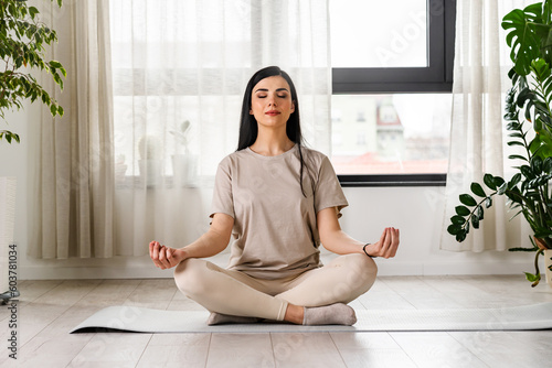 Young woman meditating sitting on a yoga mat near a big window