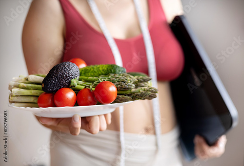 overweight woman on body composition scale,weighing balance and measure tape on neck, plate with vegetables,asparagus,tomatoes,cucumbers,zucchini.weight loss slim fit,healthy nutrition,diet concept.