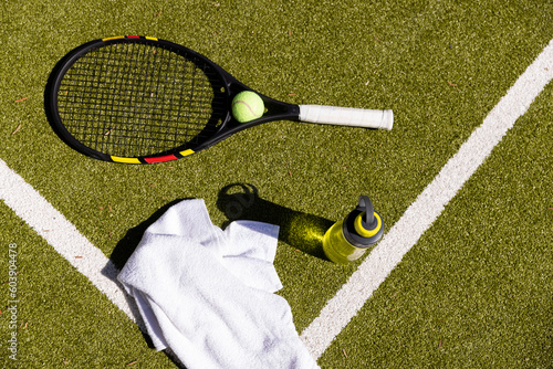 Tennis ball, racket, towel and water bottle lying on sunny outdoor grass tennis court