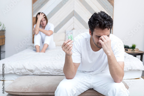 Frustrated and depressed man sitting on the edge of the bed and holding tablets for strength after conflict with his wife because of his erectile dysfunction problem
