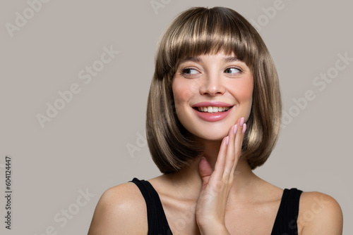 Happy young woman with bob hairstyle smiling while touching her face with her hand. Gray background
